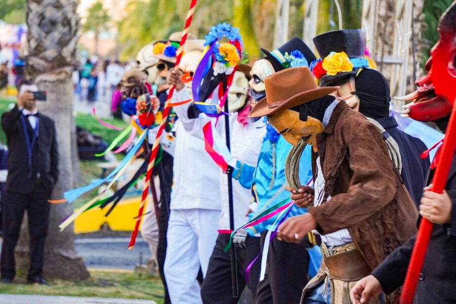 Desfile de comparsas con máscaras, sombreros y listones de colores a lo largo de una vialidad arbolada
