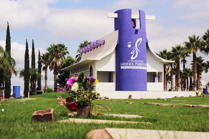 La capilla de Jardines Perpetuos, un edificio blanco con torre morada y el emblema de la marca, entre palmeras y cipreses; al frente, un arreglo de flores sobre una placa