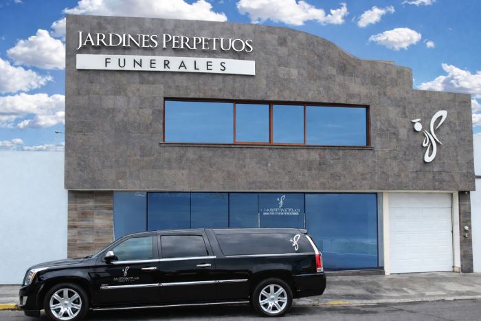 Fachada de piedra de Jardines Perpetuos Funerales con su letrero y el emblema, y una carroza fúnebre negra estacionada al frente