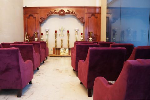 Sala de velación con sillones de terciopelo guinda frente a un altar de madera tallada con crucifijo y candeleros