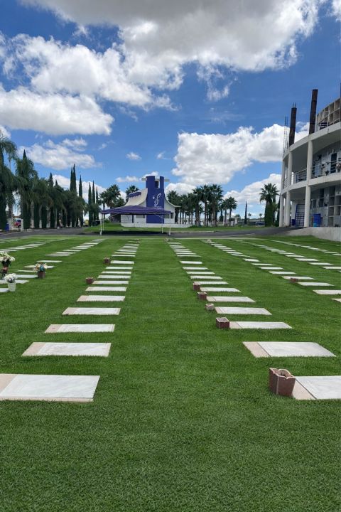 Hileras de placas conmemorativas sobre el césped, con floreros, y al fondo la capilla y el edificio de nichos